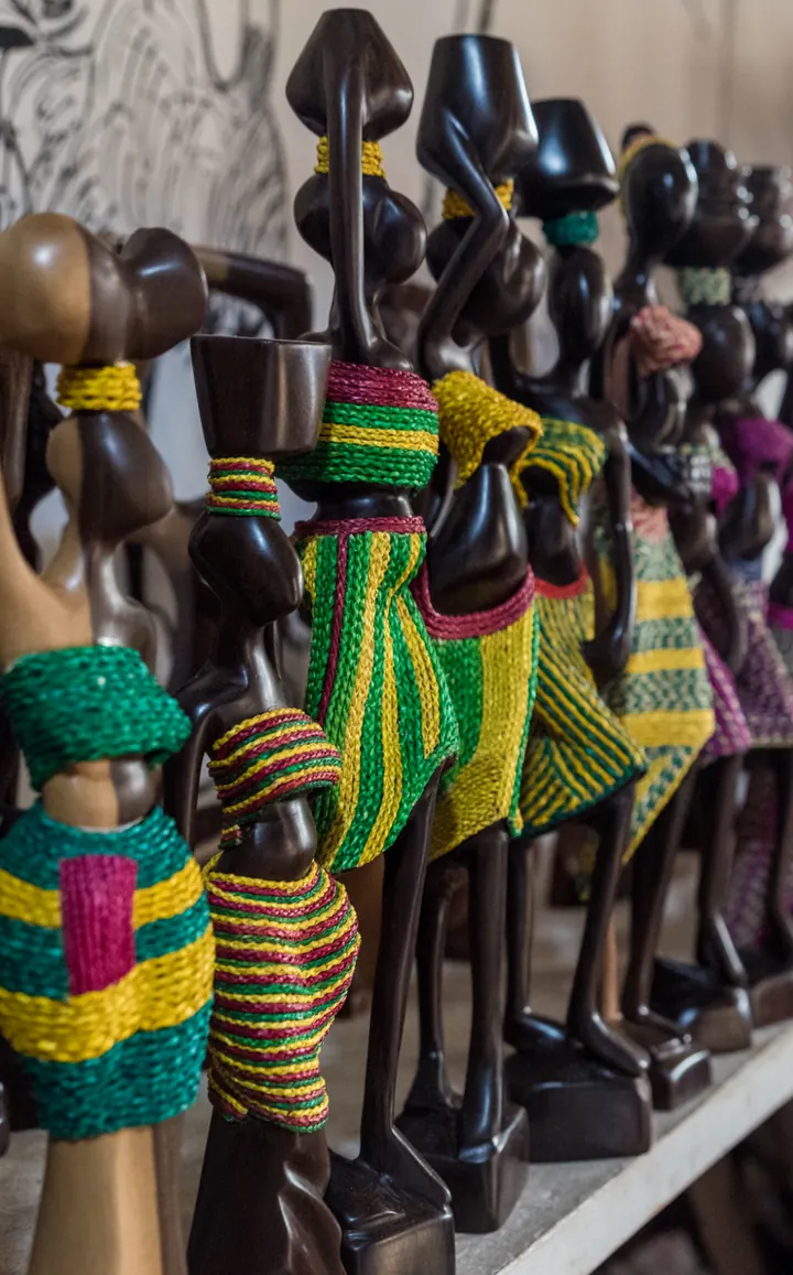 Rangée de statuettes en bois sombre habillées de bandes tissées vertes, jaunes et roses, dans une échoppe