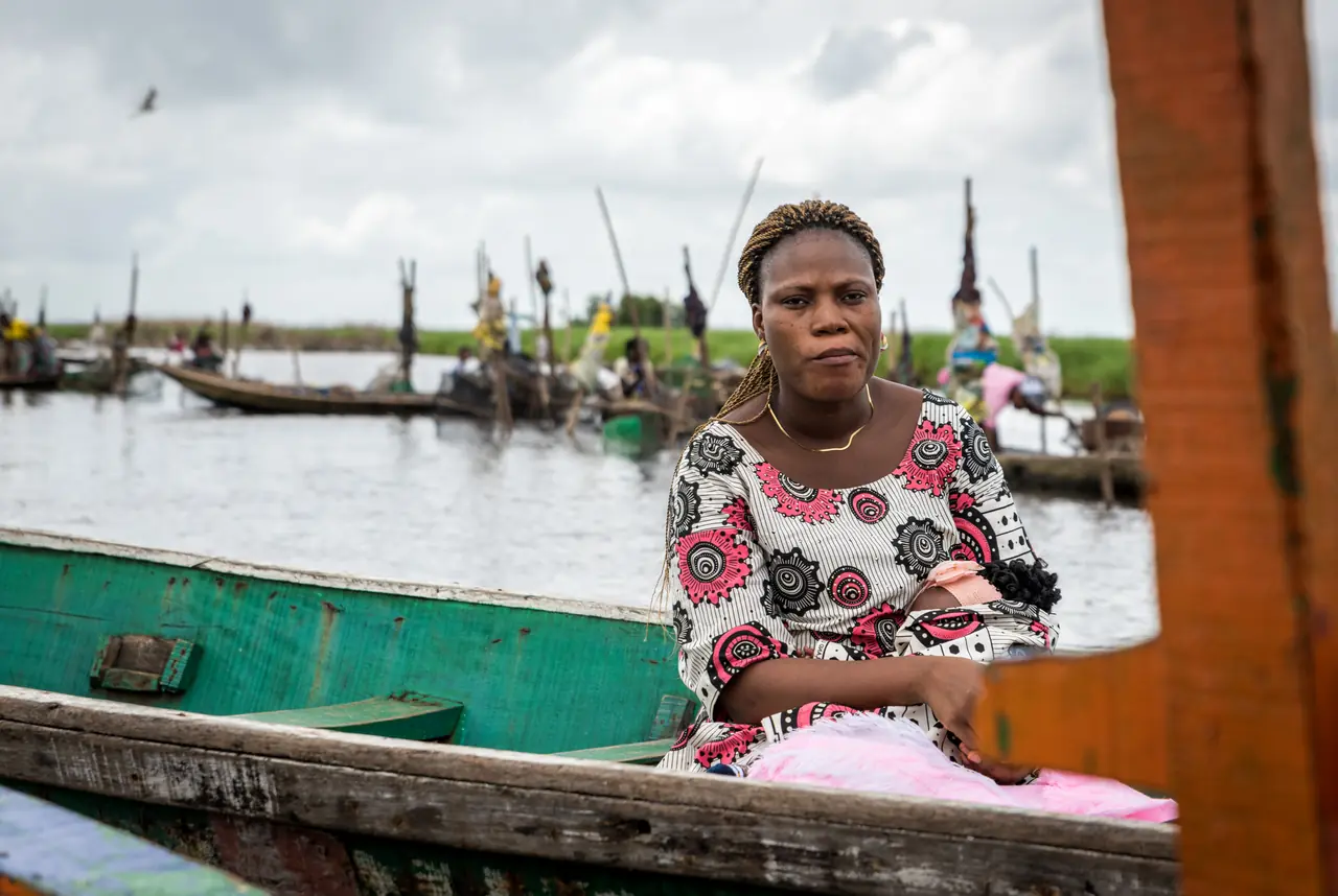 Une femme en robe à motifs assise à l’avant d’une pirogue verte sur le lac Nokoué, les pirogues du marché flottant alignées derrière elle