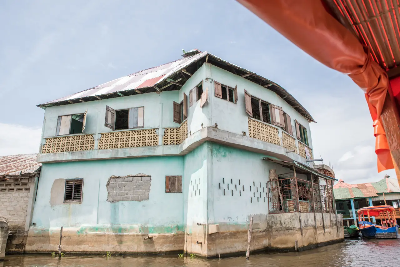 Une maison turquoise à étage bâtie sur pilotis au milieu du lac Nokoué, toit de tôle et volets de bois, vue depuis une pirogue abritée d’un taud orange