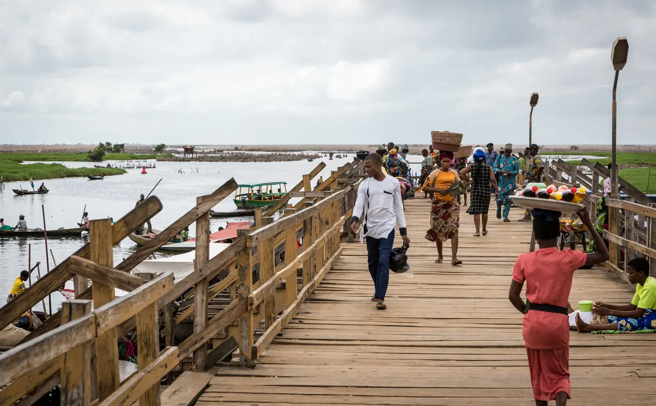 La passerelle de bois de Ganvié traversée par des habitants, une femme portant sa charge sur la tête, le lac et les toits du village de part et d’autre