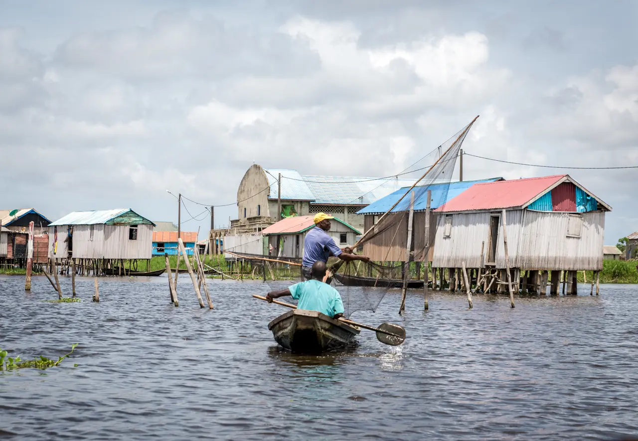 Un homme pagaie debout dans sa pirogue devant la ligne des maisons sur pilotis de Ganvié, façades bleues, vertes et ocre posées au-dessus de l’eau