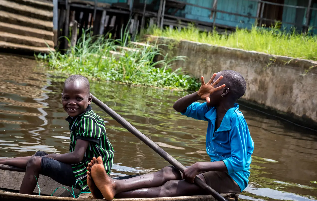 Deux piroguiers de Ganvié éclatent de rire, l’un pieds nus assis en tailleur, l’autre debout à la perche, leur pirogue longeant un mur bordé d’herbes