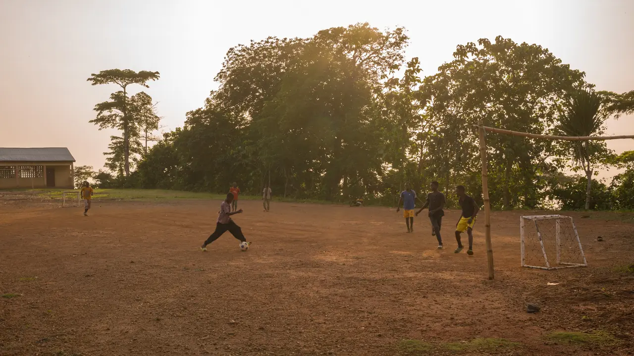Partie de football sur un terrain de terre battue au crépuscule, sous de grands fromagers, dans un village de la région de Kpalimé