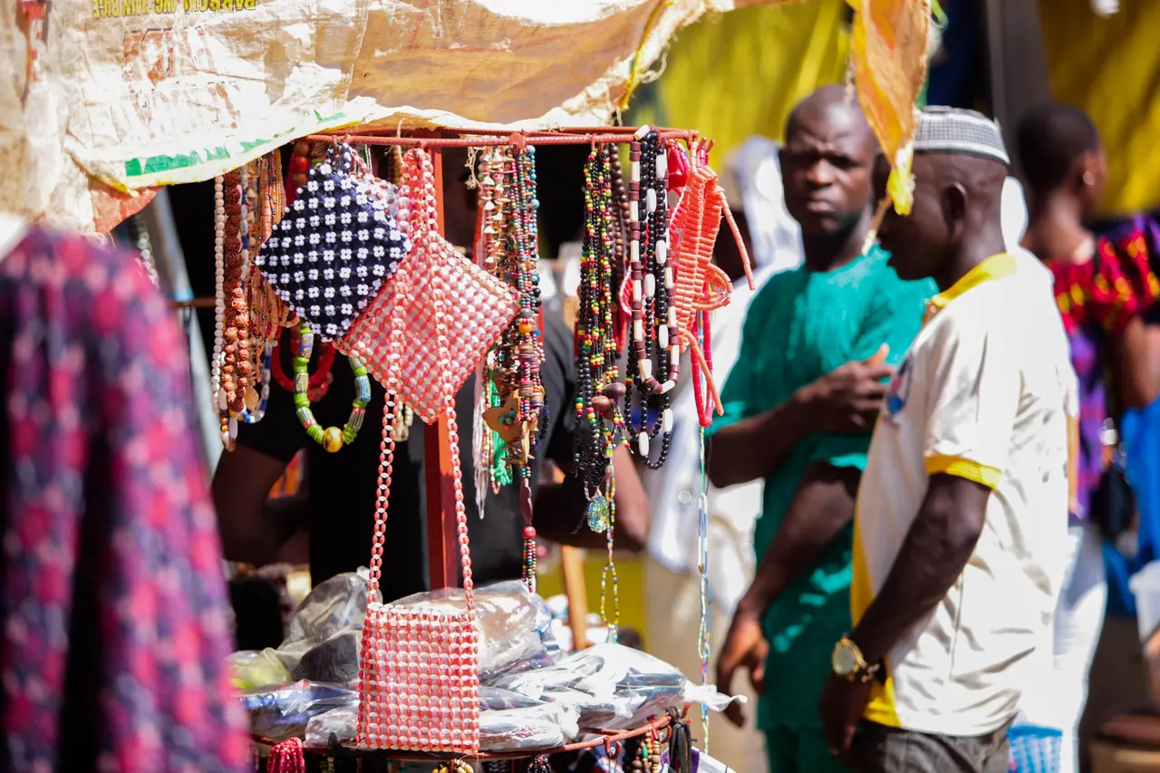 Étal d'artisanat de perles sur un marché du nord du Bénin : colliers rouges, noirs et multicolores suspendus et sacoches tressées en perles, avec des passants derrière l'étal.
