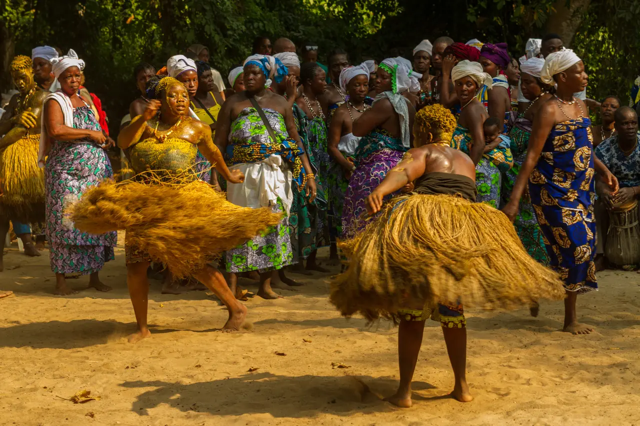 Un danseur en costume de raphia tourne sur lui-même sur la terre battue, sa jupe déployée en corolle, devant un cercle de femmes en pagnes wax