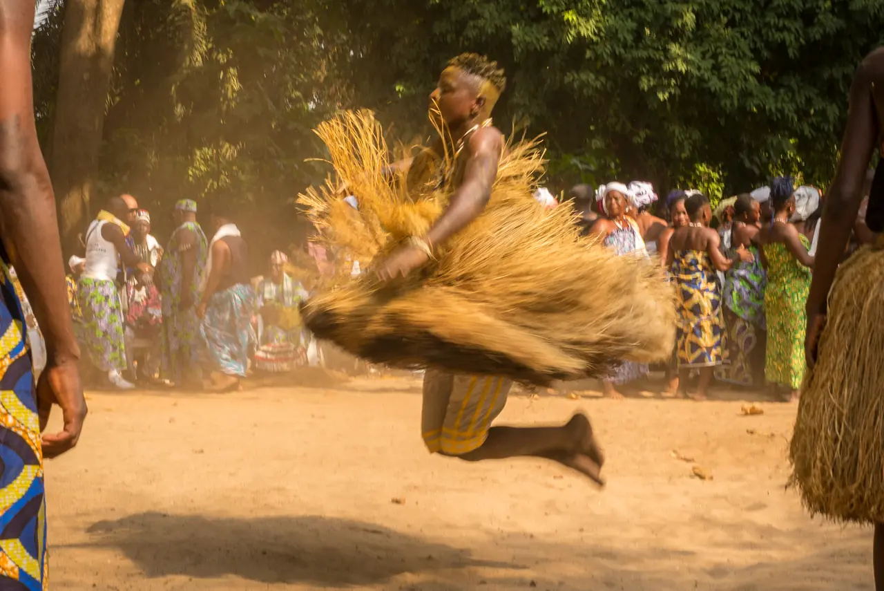Un danseur en raphia saisi en plein saut, les deux pieds décollés du sol et la jupe de fibres en suspension, la foule des Vodun Days derrière lui