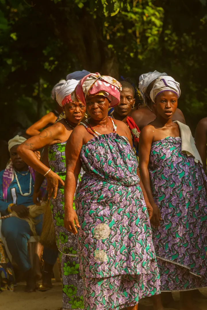 Quatre femmes en pagnes wax assortis et foulards noués se tiennent côte à côte pendant les Vodun Days, jupes de raphia des danseurs à l’arrière-plan