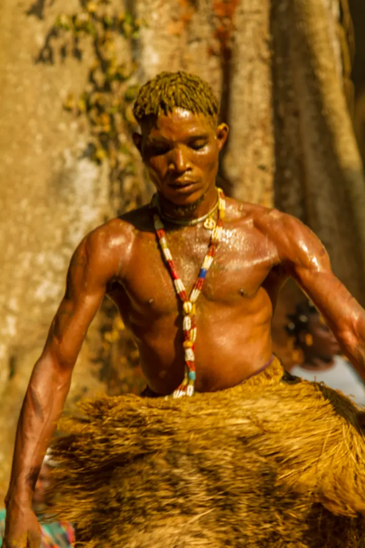 Buste d’un initié aux Vodun Days portant plusieurs colliers de perles rouges et blanches, la peau saupoudrée d’ocre sous la lumière des arbres