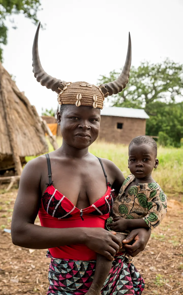Une femme coiffée d’une parure à cornes tient son enfant devant les cases du village