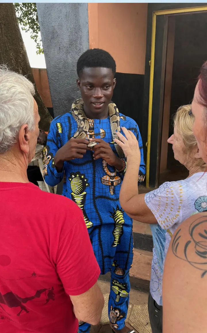 Un jeune homme en tenue wax bleue présente un python à deux voyageurs, devant le temple