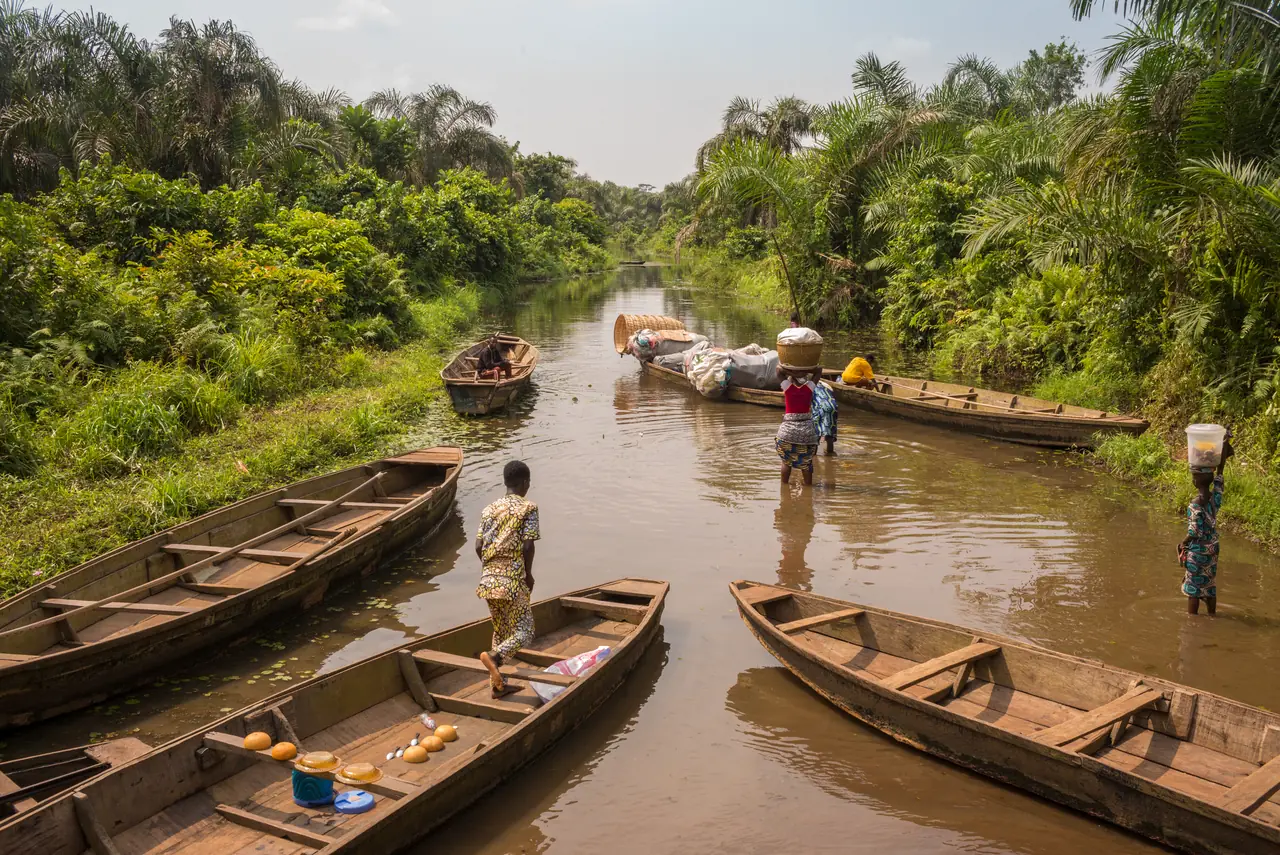 Des pirogues de bois amarrées côte à côte le long du chenal de la Rivière Noire, un homme debout dans l’une d’elles, la palmeraie fermant les deux rives