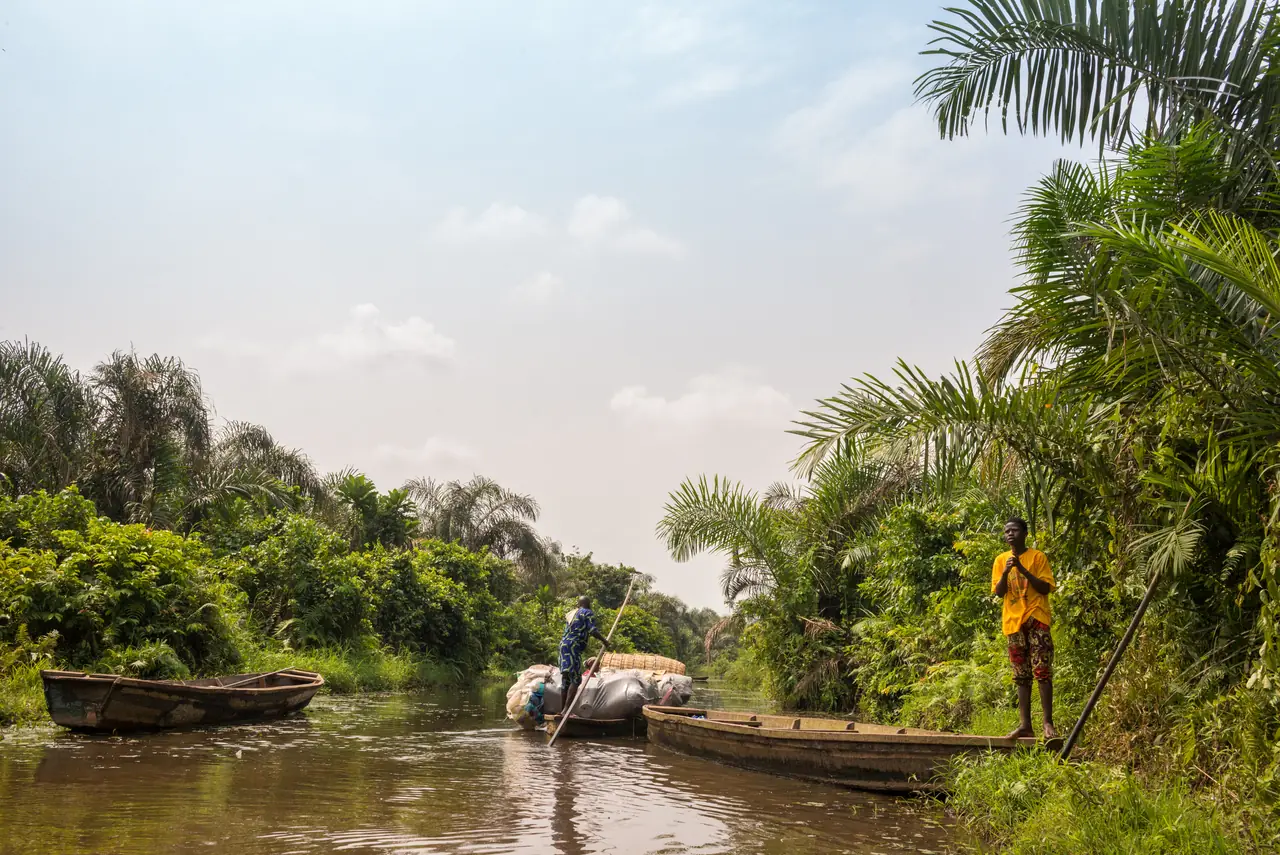 Un piroguier pousse sa barque à la perche sur l’eau plate de la Rivière Noire, deux pirogues amarrées à sa gauche et un mur de palmiers derrière lui