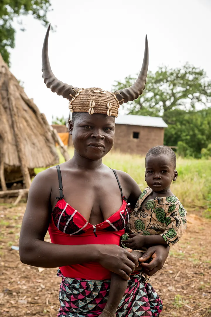 Une femme du pays Tamberma coiffée de cornes et de cowries porte son enfant sur la hanche devant les maisons de terre du village